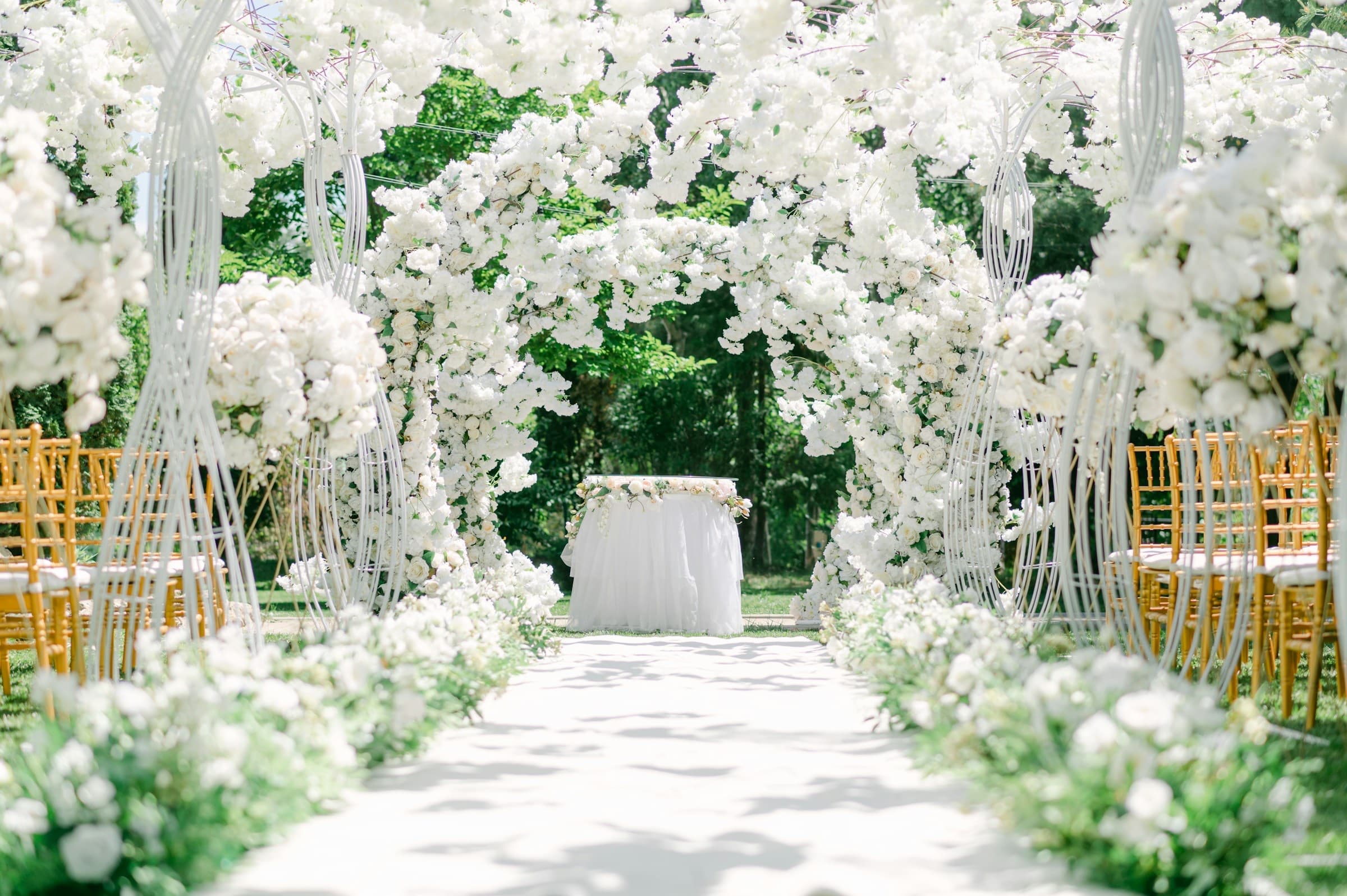 A white floral archway at the end of a garden aisle lined with gold chairs